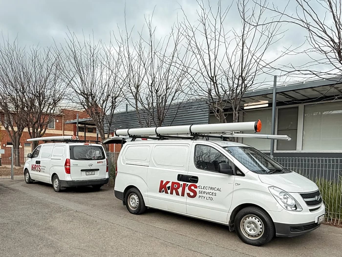 Two Kris Electrical Services vans with roof racks parked outside a commercial building